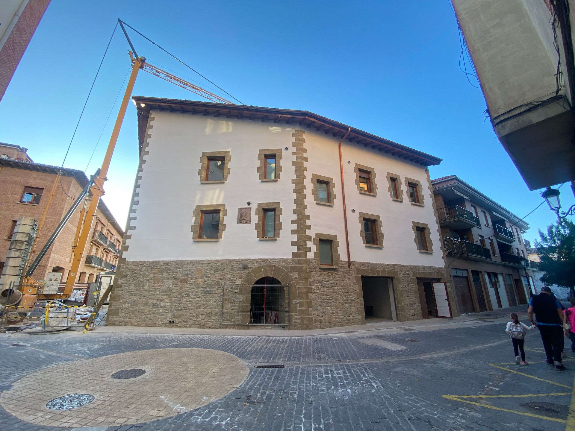 Edificio en construcción; grúa presente. Base de piedra, pisos superiores de color claro. Calle adoquinada.