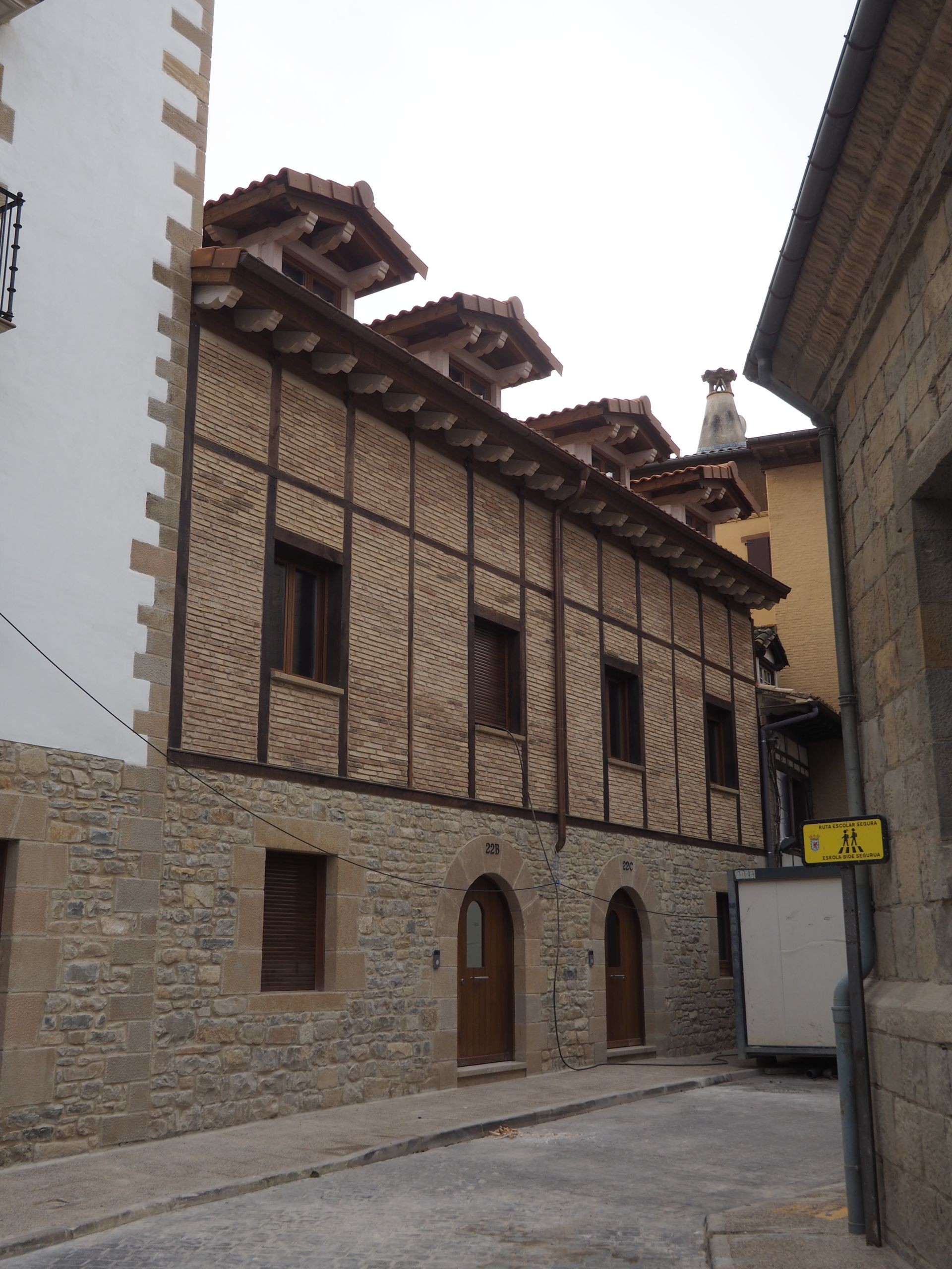 Edificio de dos plantas con base de piedra, vigas de madera y buhardillas con techo marrón en una calle estrecha.