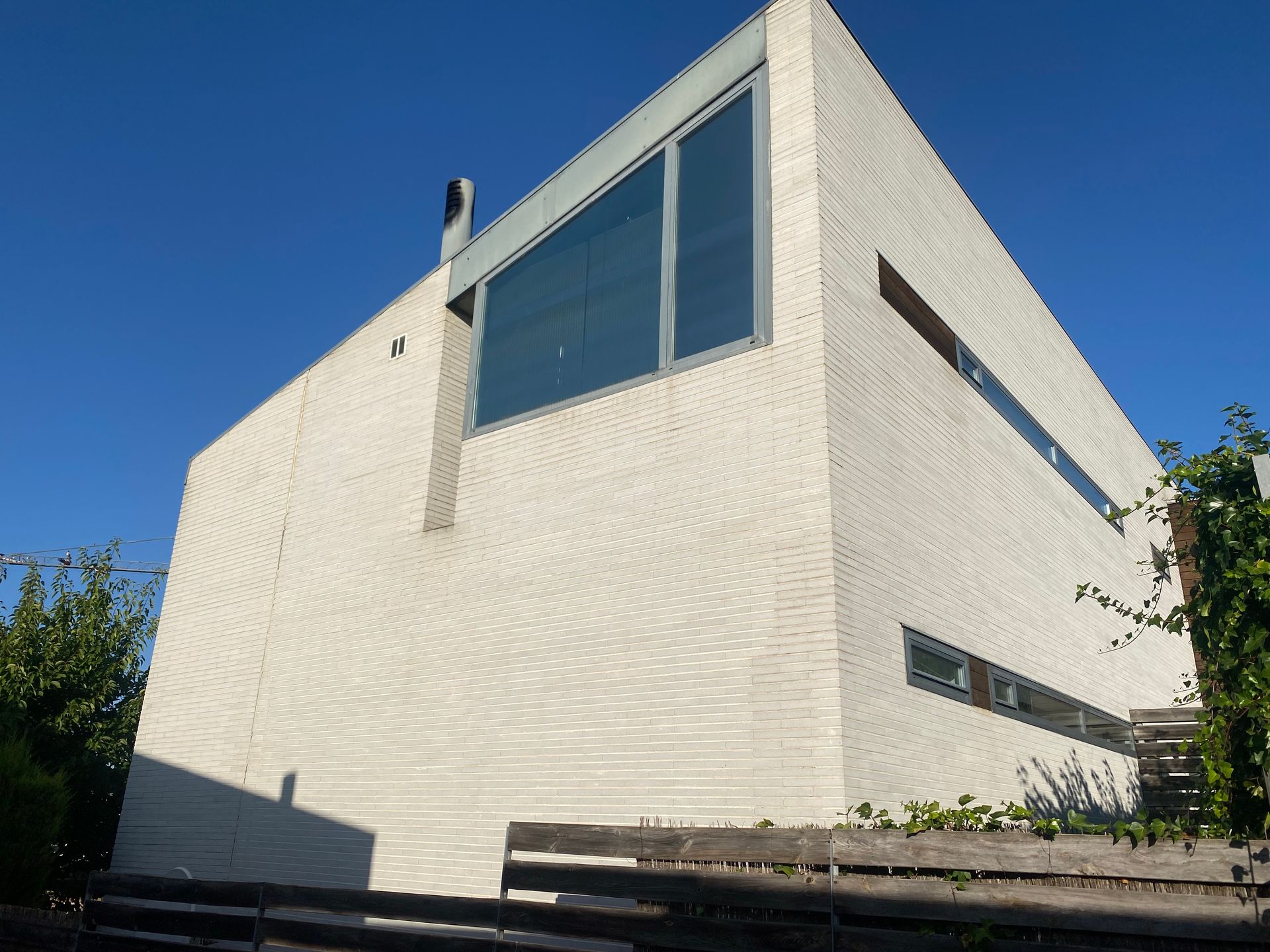 Edificio moderno blanco con ventanal, pequeñas ventanas rectangulares, chimenea y cielo azul.