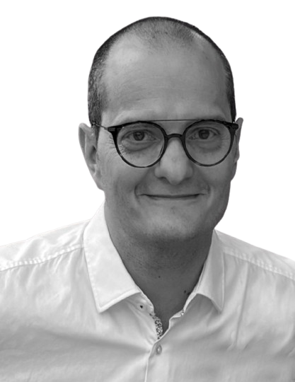 Un hombre con gafas y camisa abotonada sonríe, mirando directamente al espectador, sobre un fondo blanco.