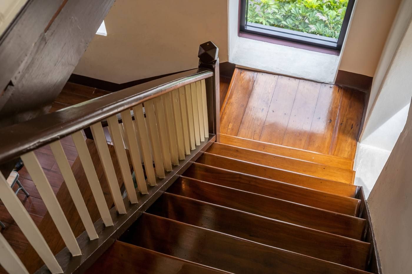 Escalera de madera con barandilla que baja hasta una puerta luminosa y vegetación en el exterior.