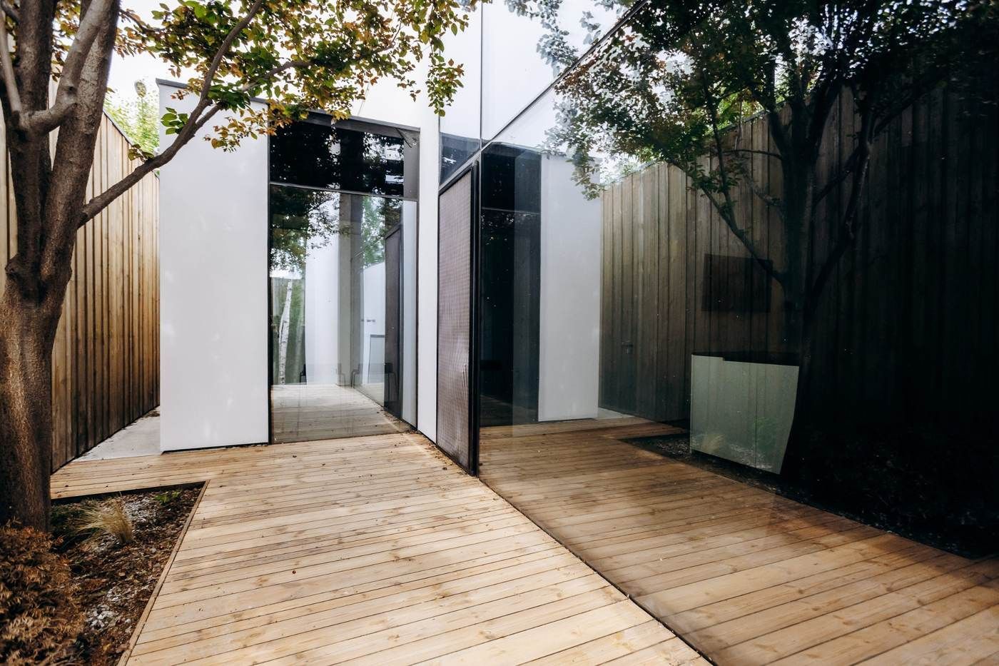 Entrada moderna a la casa con puertas de cristal, paneles blancos y una terraza de madera rodeada de árboles y vallas.