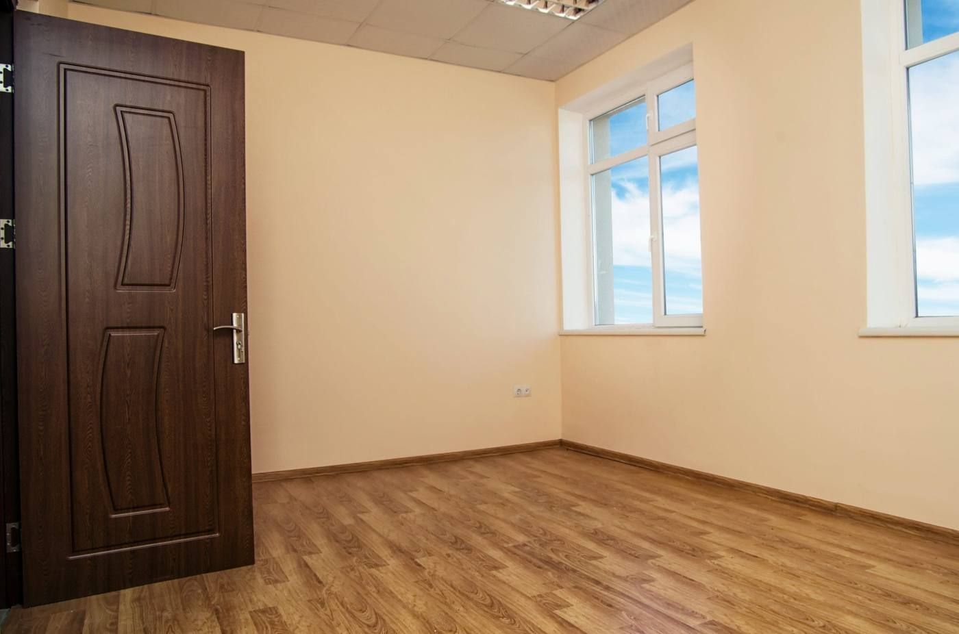 Habitación vacía con suelo de madera, paredes color crema, una puerta de madera oscura y dos ventanas con vistas al cielo.