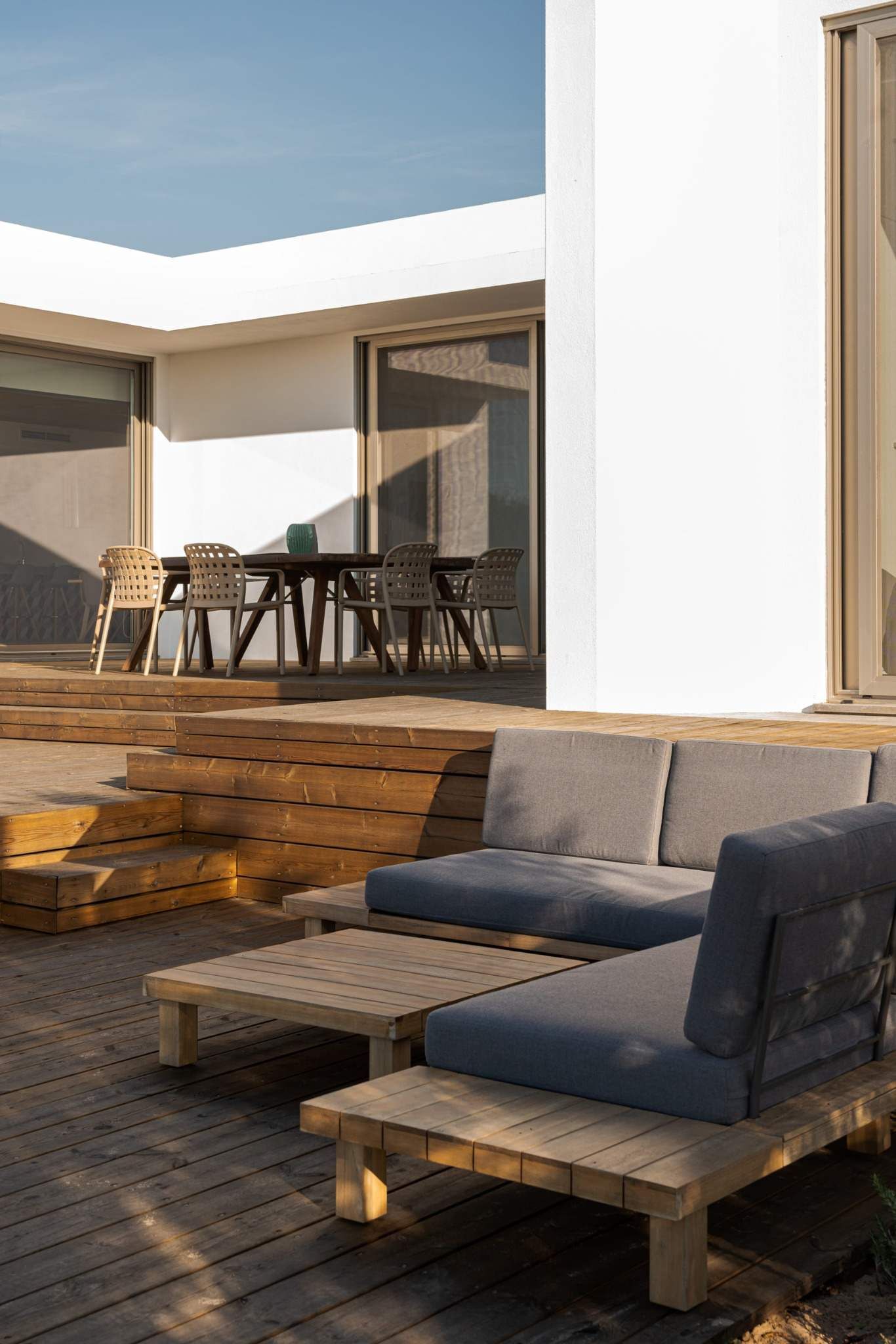 Patio moderno con terraza de madera, tumbonas de mimbre, mesa de comedor y paredes de estuco blanco.