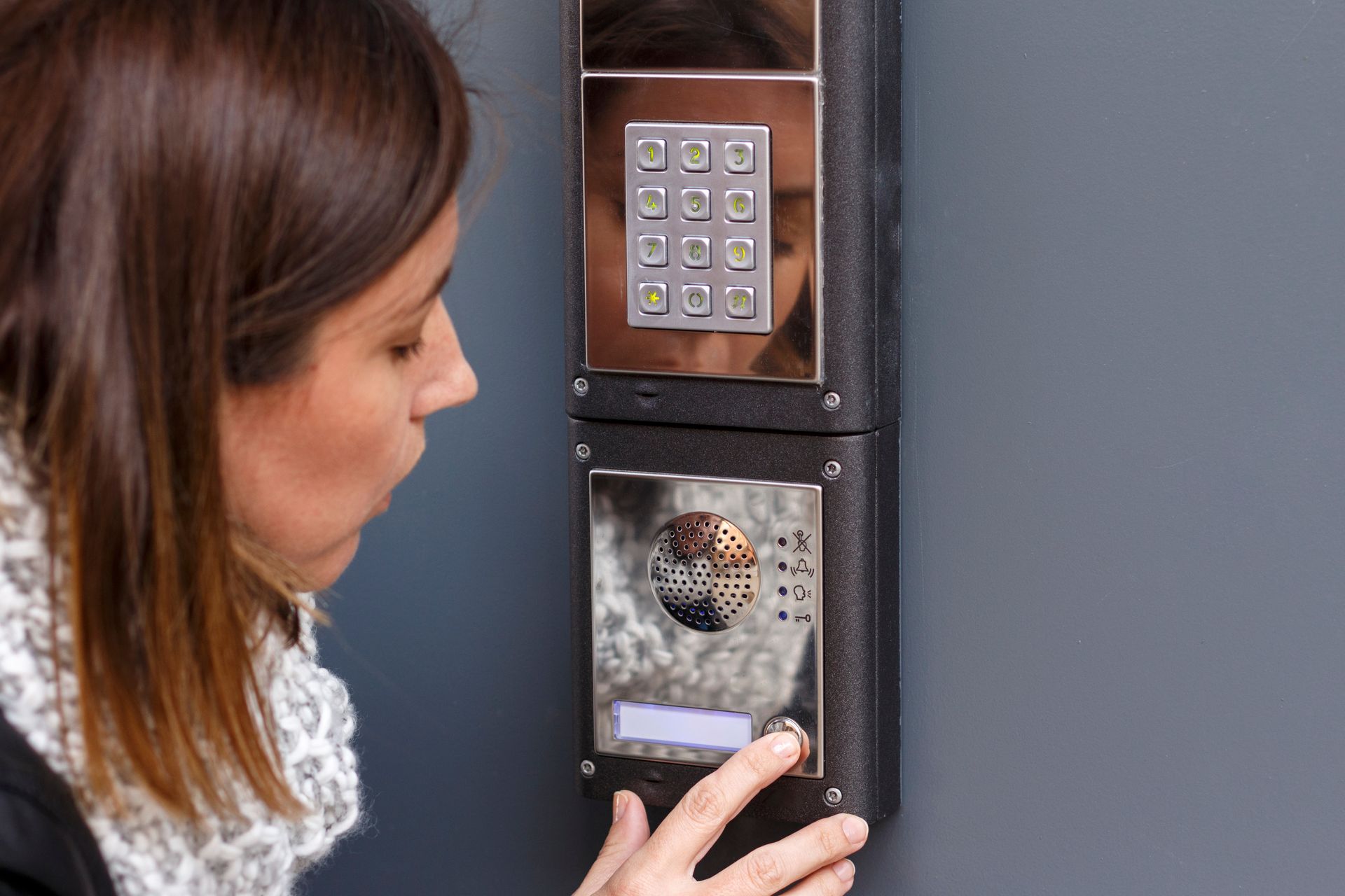 Une femme utilise un système d'interphone dans un immeuble ; elle appuie sur un bouton et regarde l'appareil sur un mur gris.