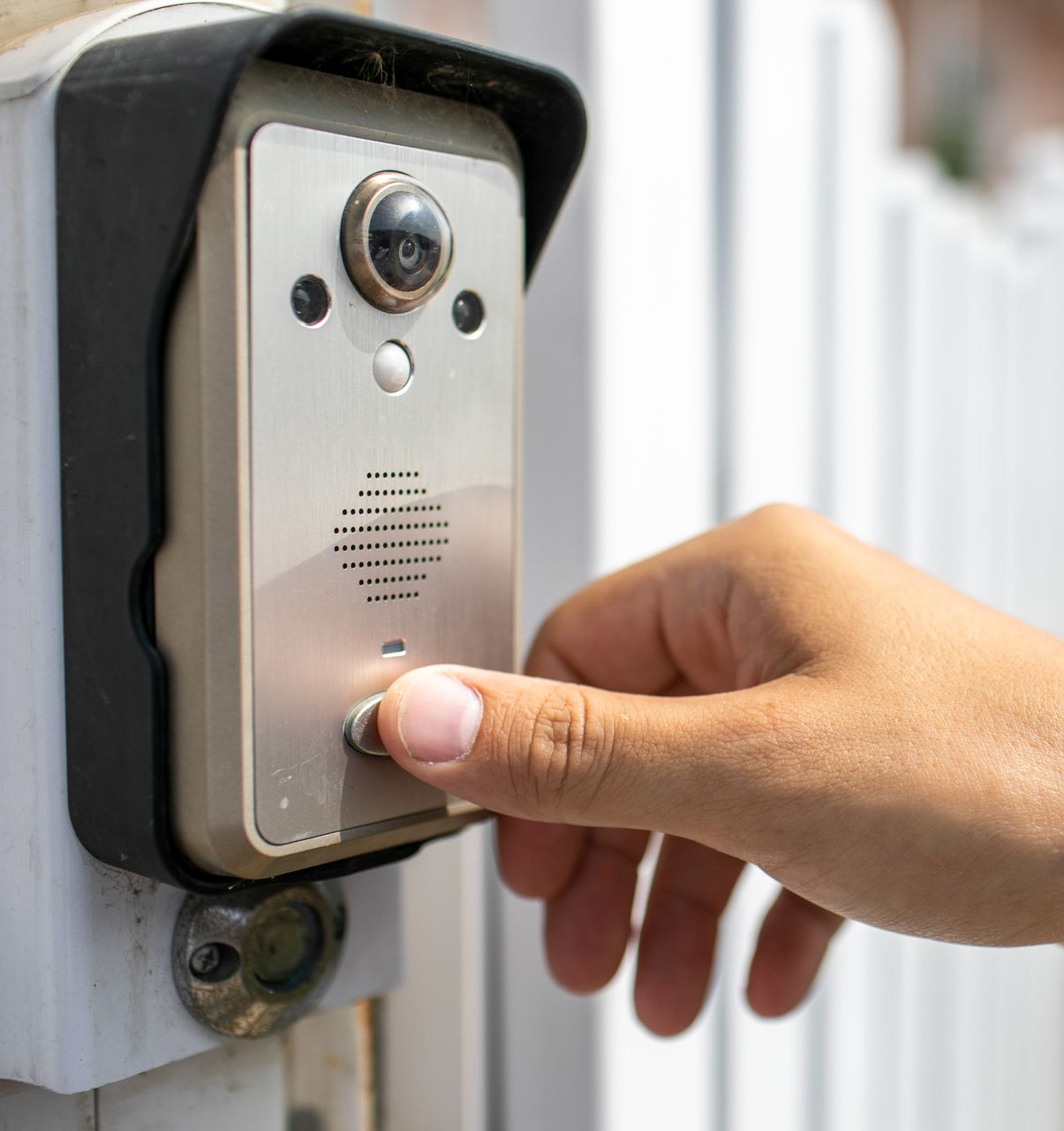 Main appuyant sur un bouton de sonnette sur un système d'interphone argenté et noir monté sur un portail blanc.