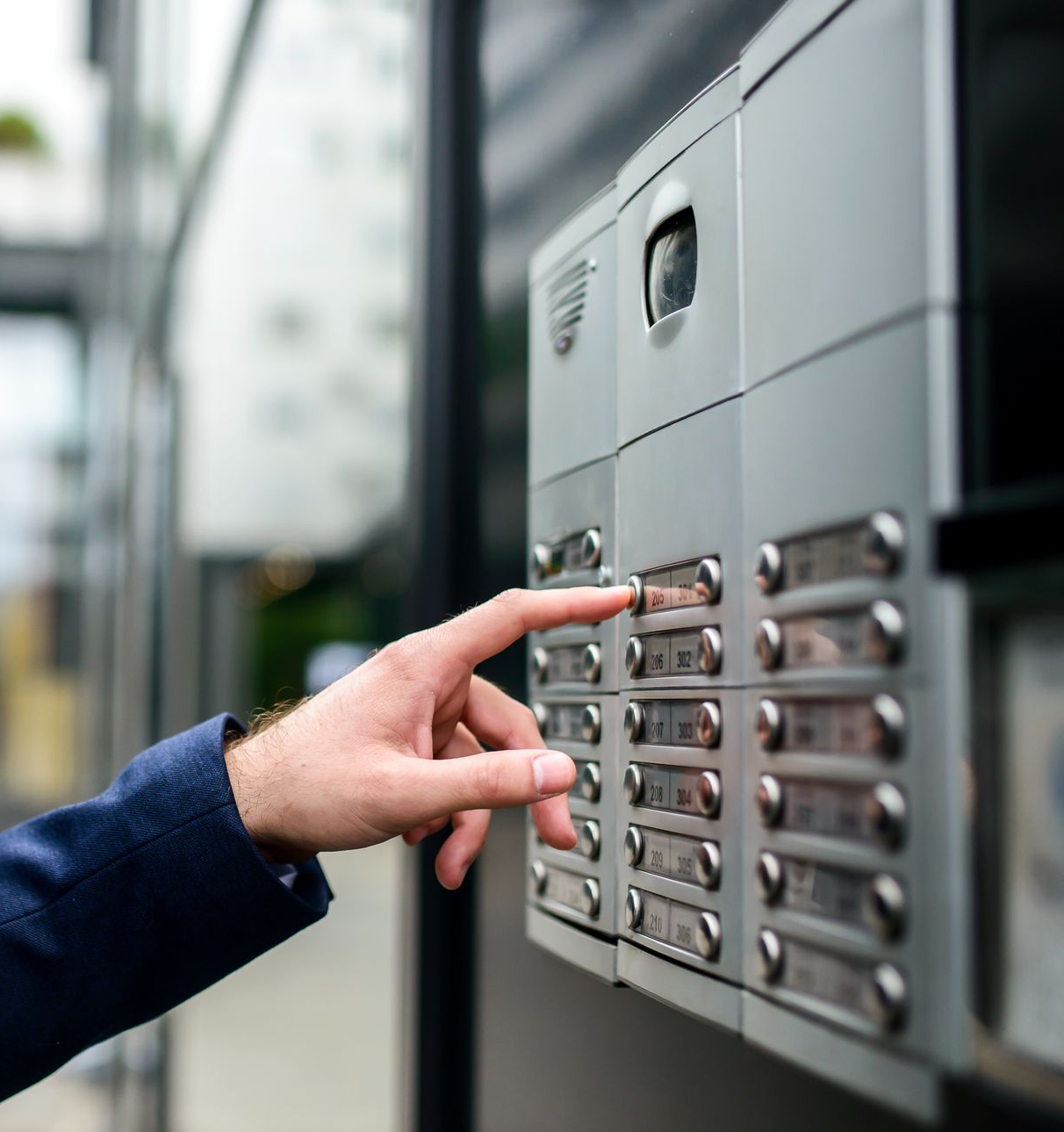 Main appuyant sur un bouton d'un panneau d'interphone argenté près d'un bâtiment.