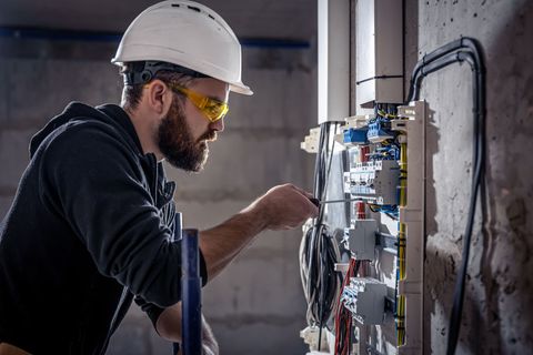 Électricien portant un casque et des lunettes de sécurité travaillant sur un boîtier de circuit à l'intérieur.
