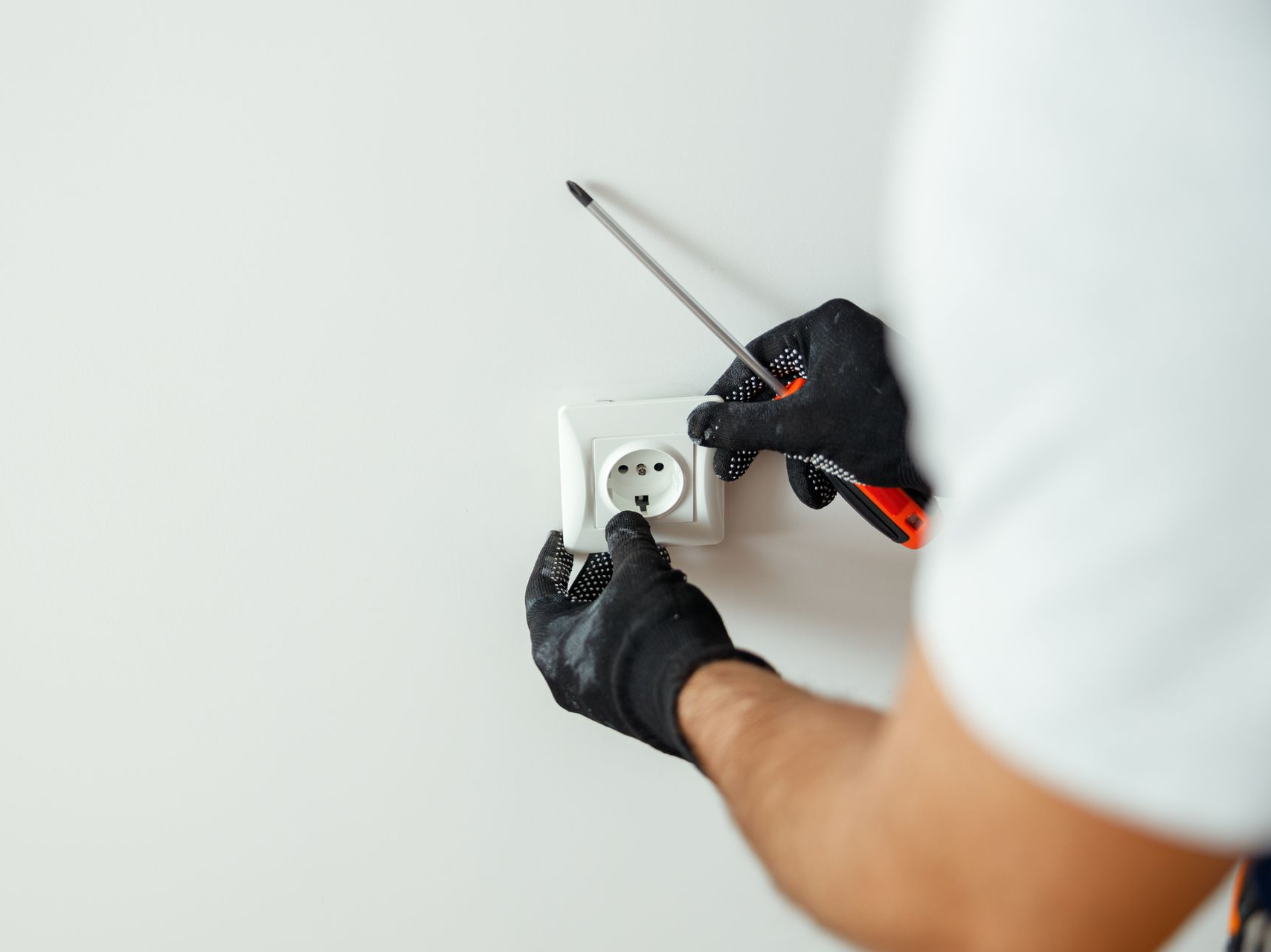 Électricien avec gants noirs utilisant un tournevis pour installer une prise électrique sur un mur blanc.