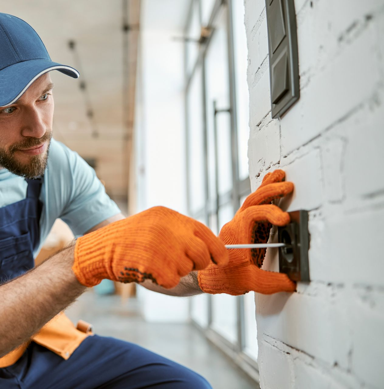 Électricien portant des gants orange installant un interrupteur sur un mur de briques blanches.