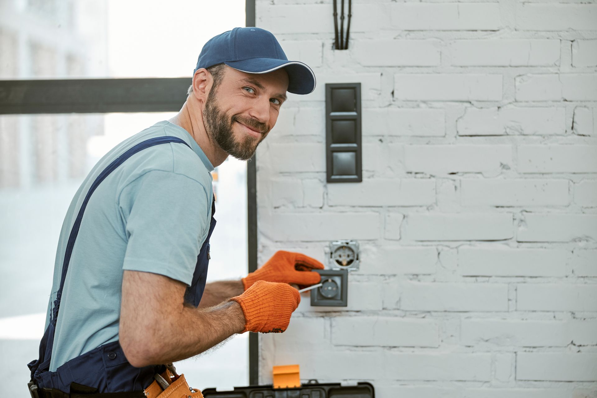 Électricien souriant tout en travaillant sur une prise, portant une casquette bleue et des gants orange dans une pièce en briques blanches.