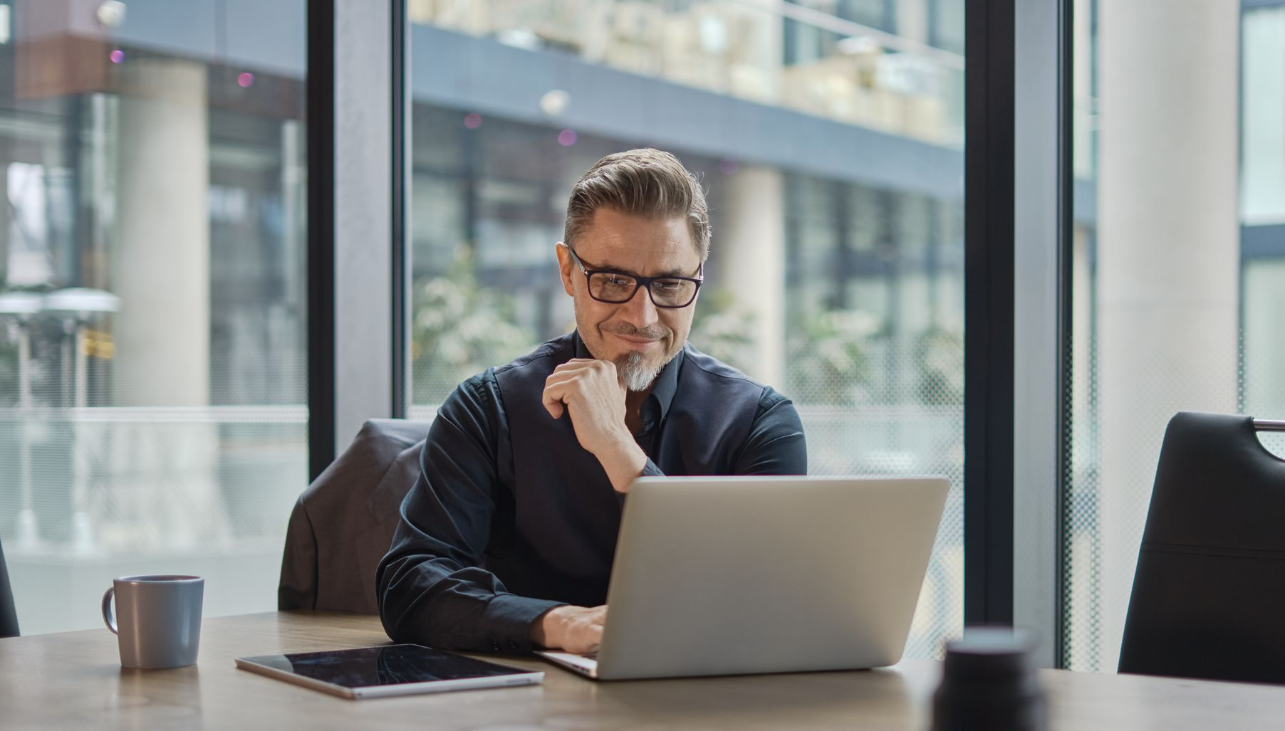 Un homme portant des lunettes souriant tout en travaillant sur un ordinateur portable dans un bureau.