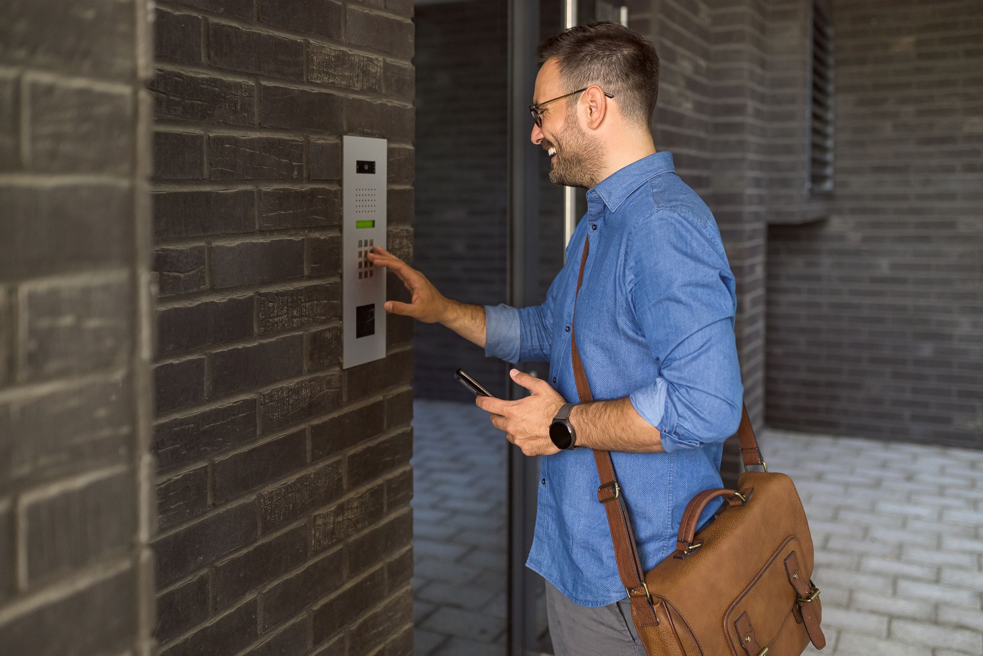 Un homme appuie sur le bouton de l'interphone, souriant, tenant un téléphone et un sac, devant un bâtiment en briques.