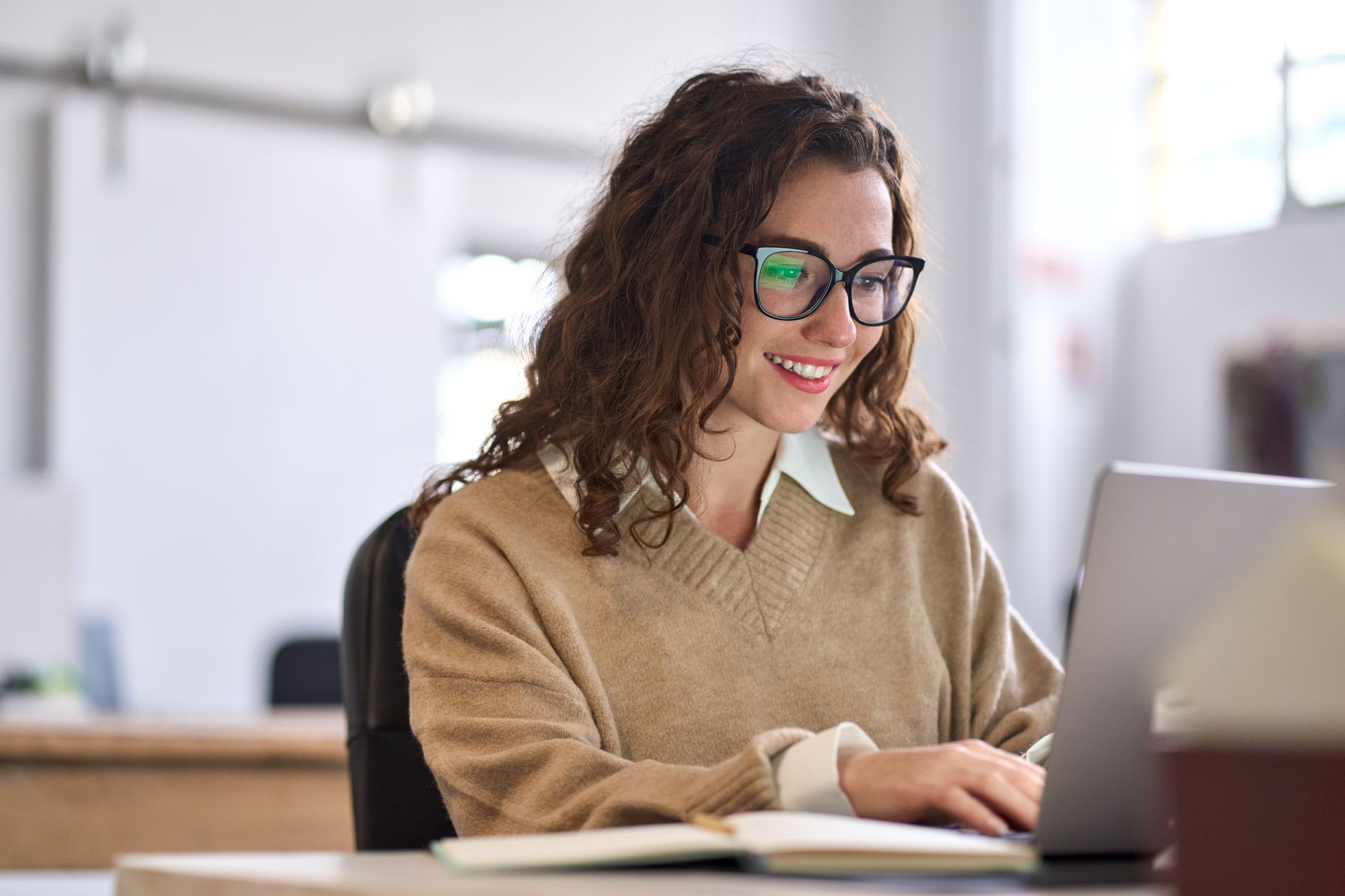 Une femme portant des lunettes sourit tout en travaillant sur un ordinateur portable dans un bureau.