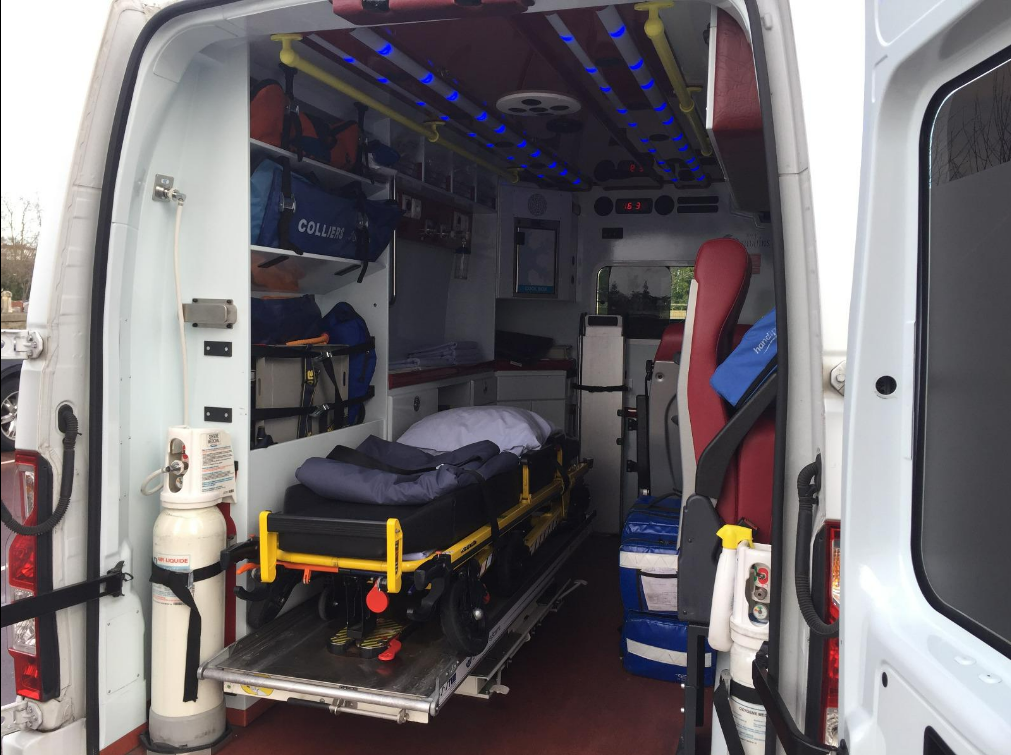 À l'intérieur d'une ambulance, on aperçoit une civière, du matériel médical et des rangements. Les portes arrière sont ouvertes.