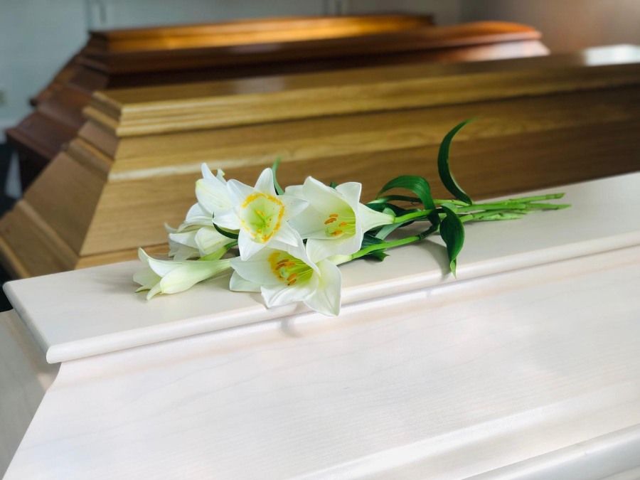 weiße Lilien auf einem weißen Sarg, im Hintergrund zwei Särge aus Holz