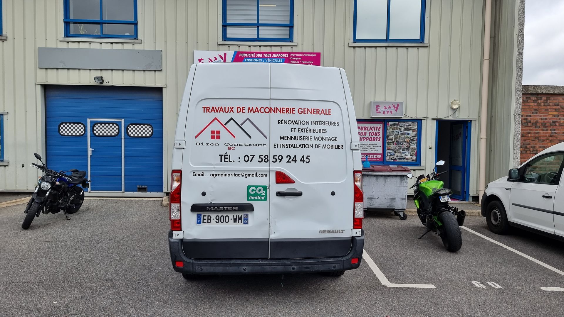 Vue arrière d'une camionnette blanche garée devant un bâtiment. Deux motos sont garées à gauche et à droite.