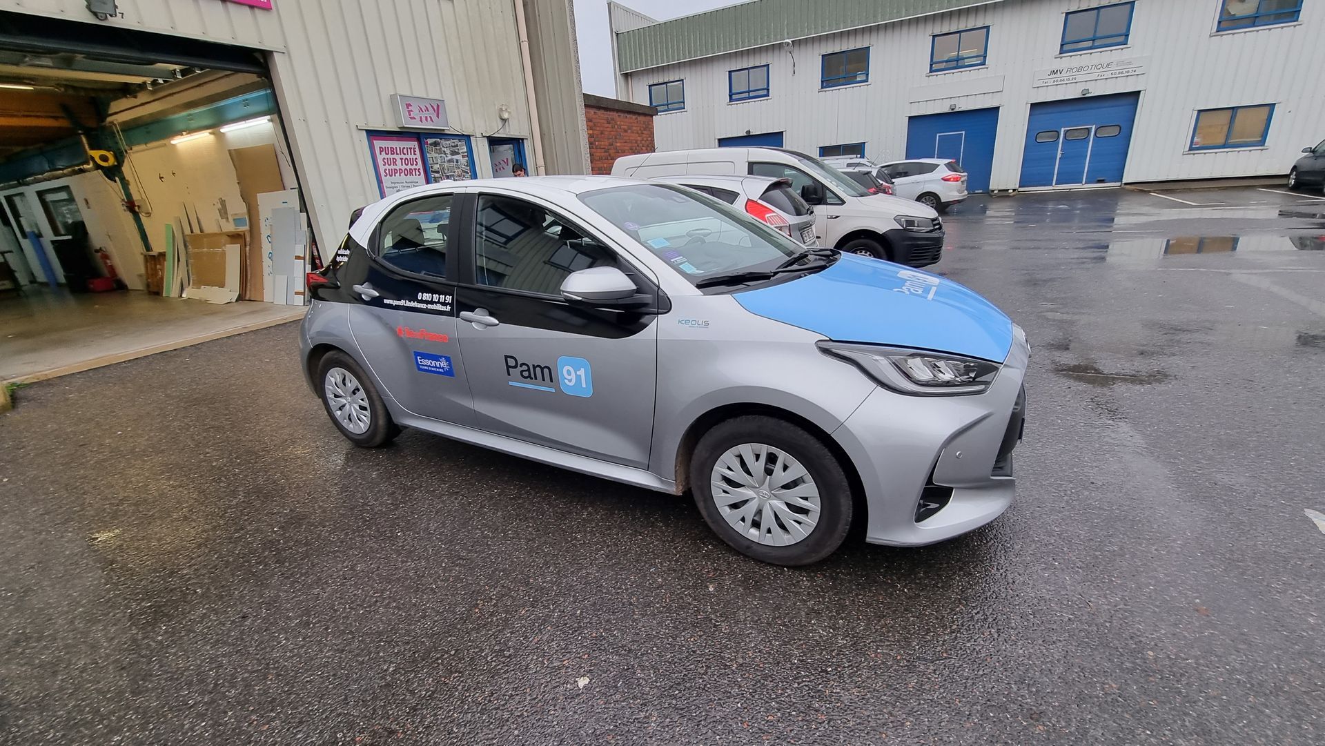 Une Toyota Yaris argentée avec capot et logos latéraux bleus est garée devant un bâtiment.