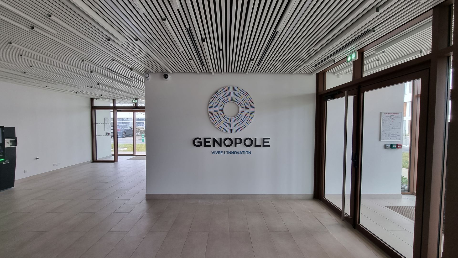 Intérieur du bâtiment GENOPOLE, avec logo sur mur blanc, sol et plafond en bois, portes vitrées.