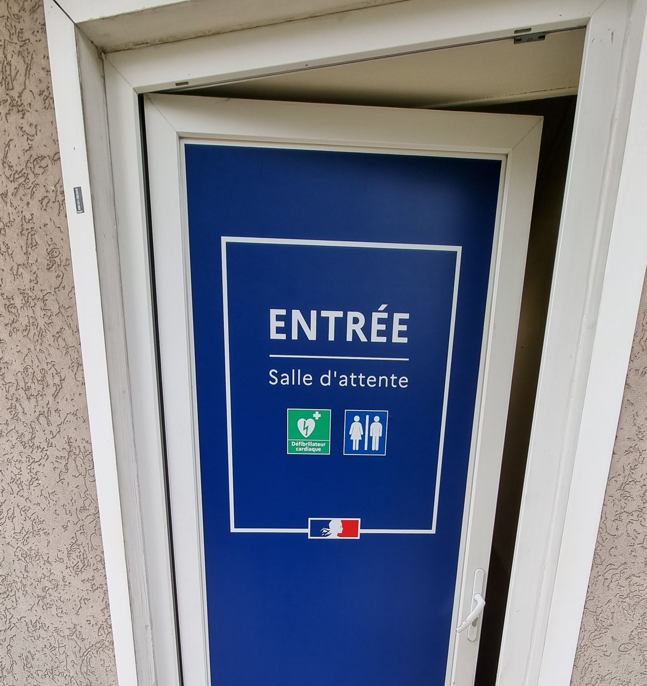 Porte bleue avec panneau ENTRÉE Salle d'attente, ainsi que les symboles de sortie de secours et de toilettes. Drapeau français en bas.