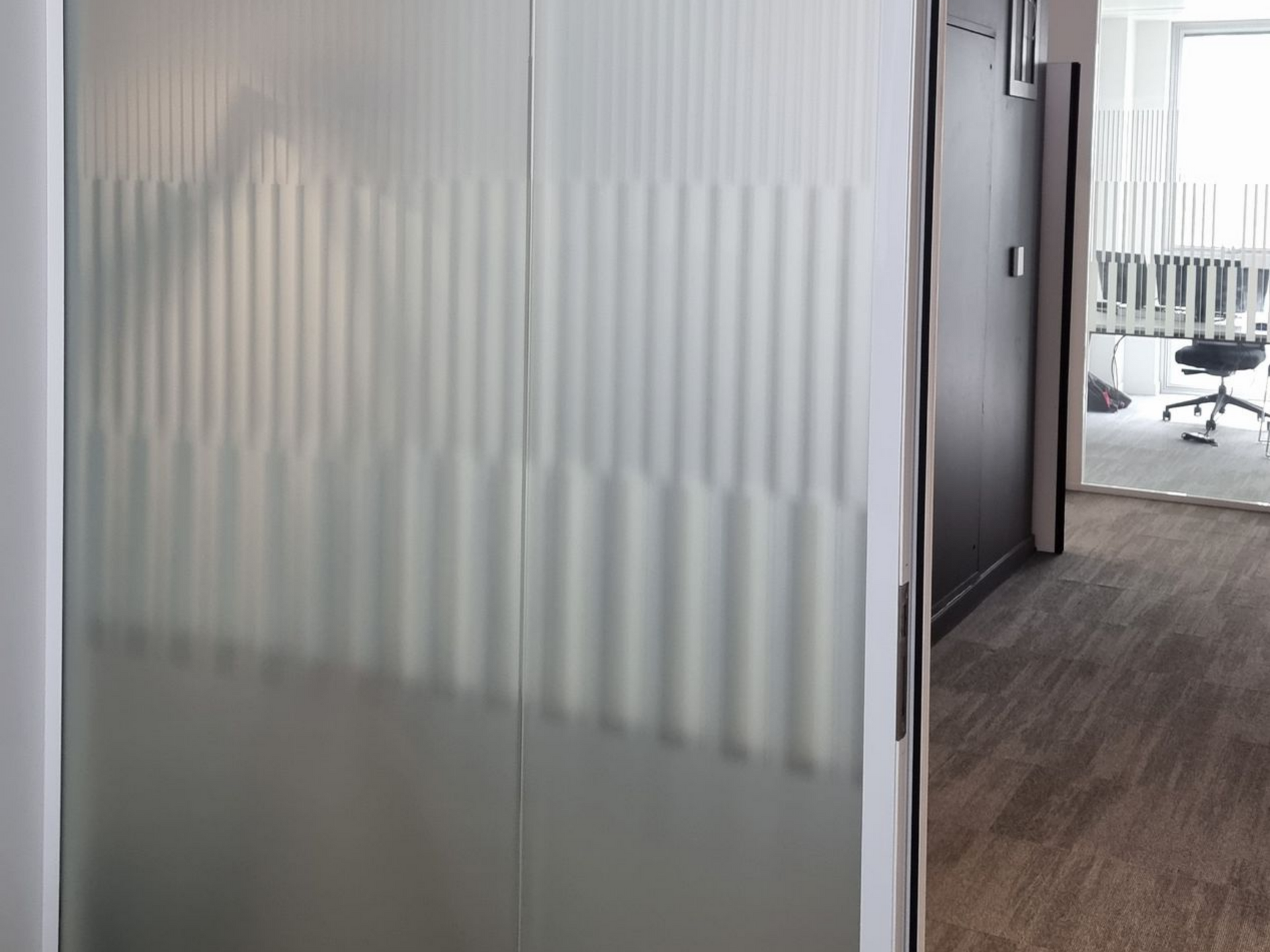 Portes coulissantes en verre dépoli à motif vertical, donnant sur un espace de bureau avec porte foncée et moquette.