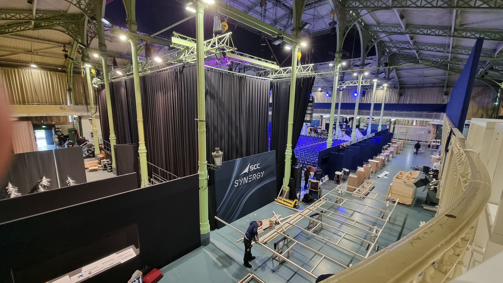 Aménagement d'événement : long hall avec rideaux noirs, éléments de scène et personnel. Éclairage au plafond et poutres métalliques.