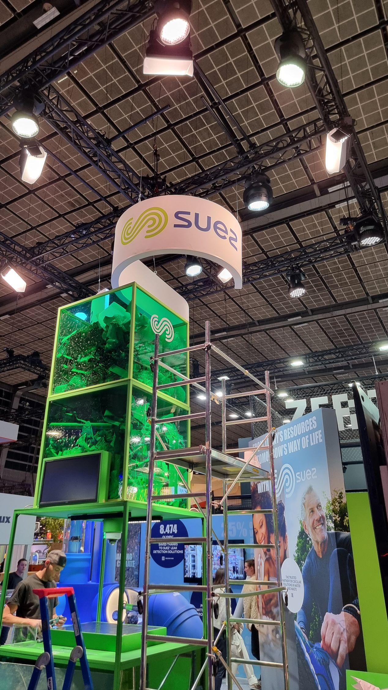 Stand de Suez, structure verte et bleue avec échafaudages, enseigne et éclairage au plafond.