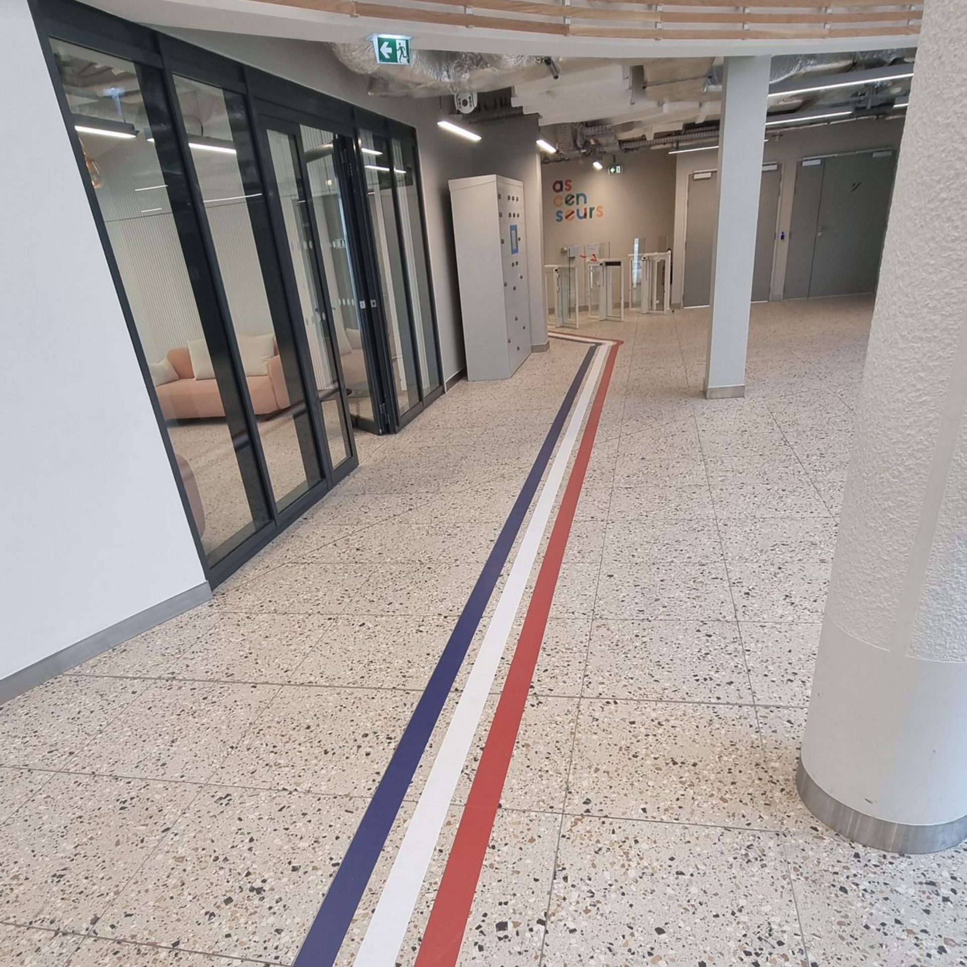 Couloir avec des rayures bleues, blanches et rouges au sol, des portes vitrées et un sol clair moucheté.