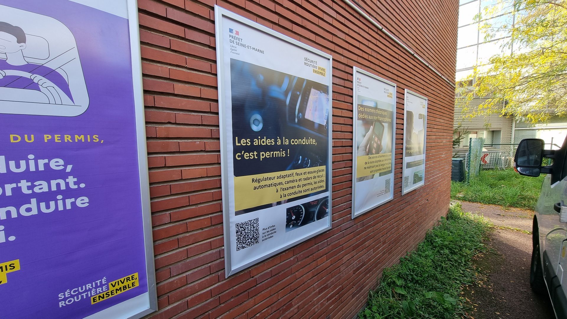 Des affiches sur un mur de briques, faisant la promotion de la sécurité routière. Texte et graphismes jaunes, blancs, violets et noirs.