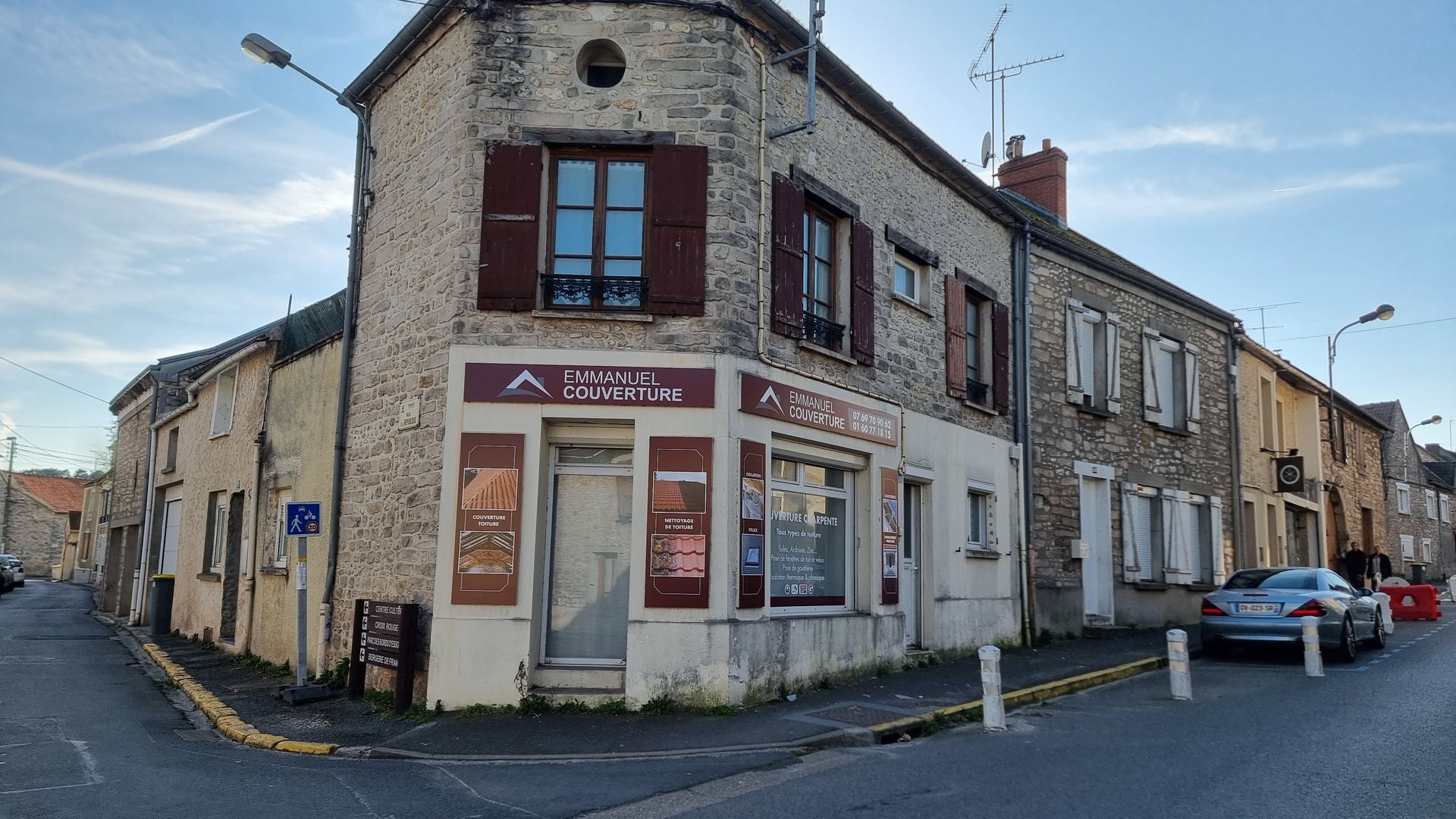 Immeuble d'angle avec magasin, façade en pierre, volets rouges, ciel bleu, voiture garée dans la rue.