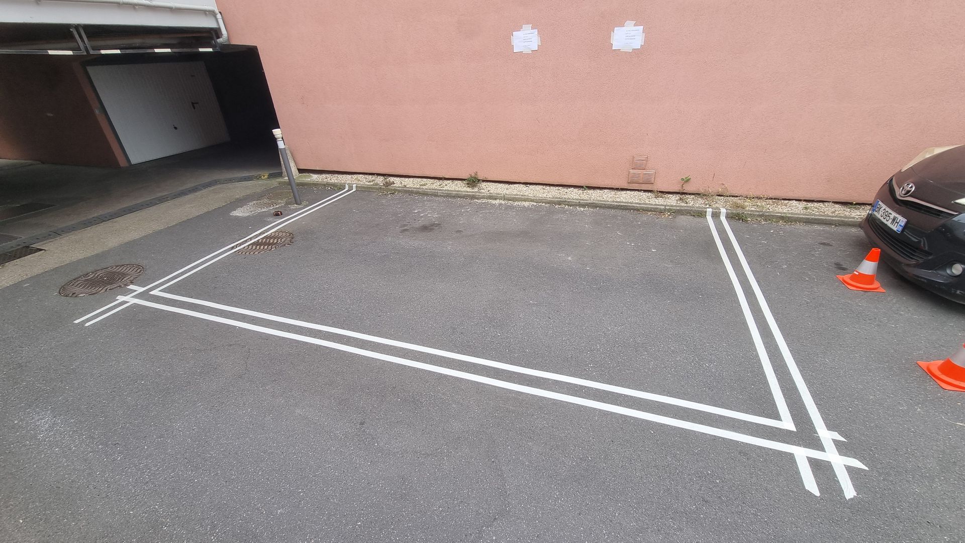 Place de stationnement délimitée par des lignes blanches sur l'asphalte, à côté d'un bâtiment et d'une voiture. Présence de cônes orange.