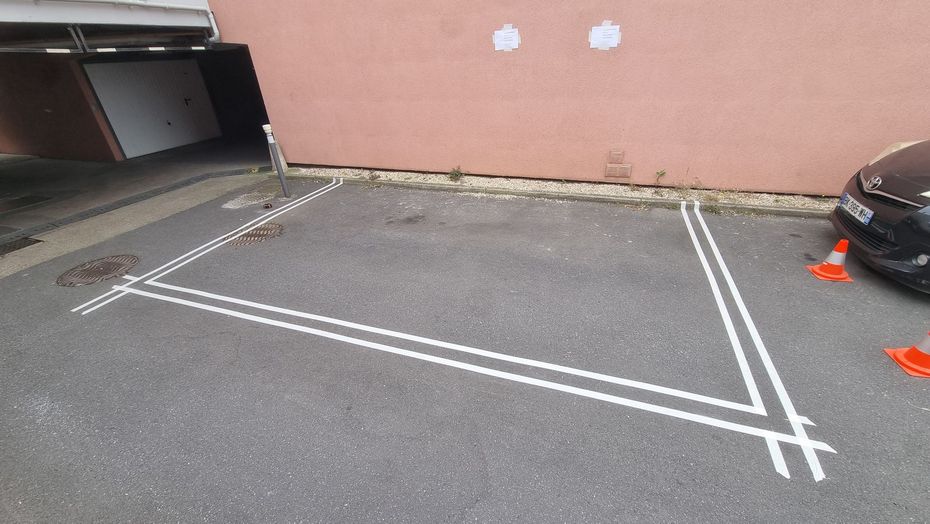 Place de stationnement délimitée par des lignes blanches sur l'asphalte, à côté d'un bâtiment et d'une voiture. Présence de cônes orange.