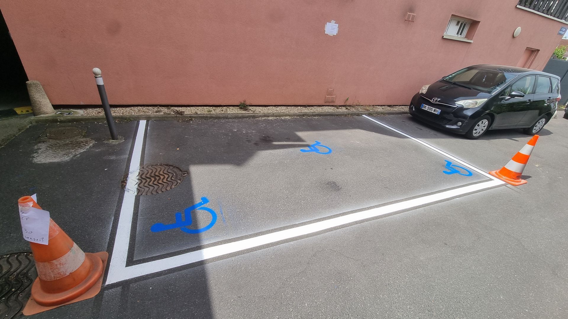 Place de parking réservée aux personnes handicapées avec symbole de fauteuil roulant bleu, cônes orange et voiture noire stationnée.