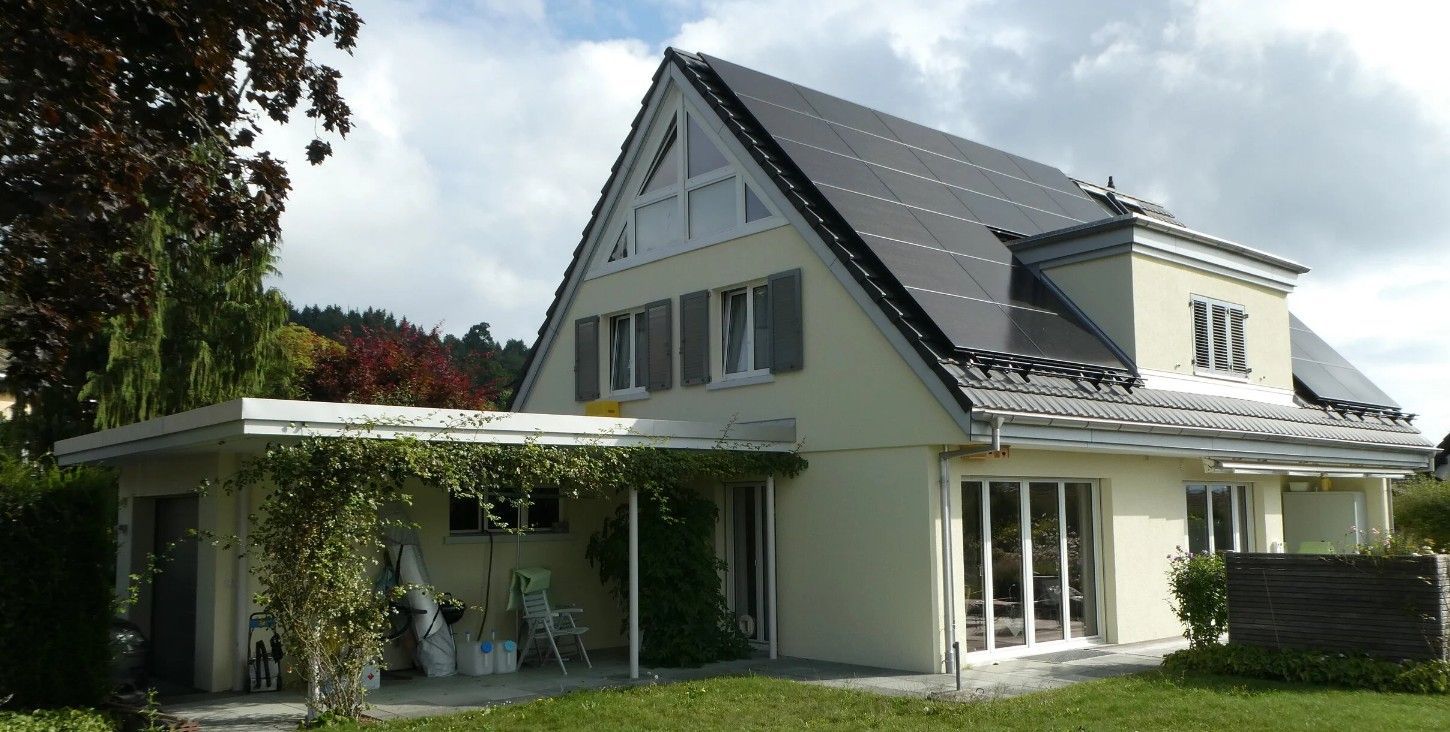 Modernes zweistöckiges Haus mit dunklem Dach, weissen Wänden und überdachter Terrasse in einem grünen Garten