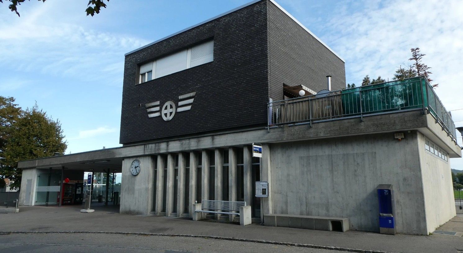 Modernes Gebäude mit dunkler oberer Fassade und geflügeltem Logo über einem unteren Betongeschoss und Säulen.