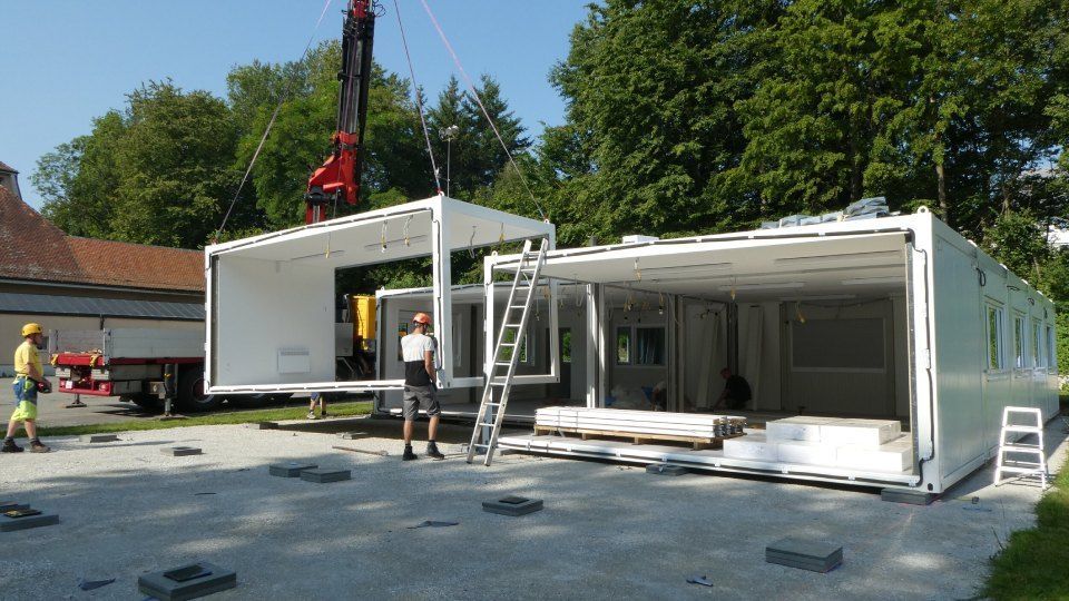 Arbeiter montieren mit einem Kran weisse Modulgebäude auf einem sonnigen Freigelände