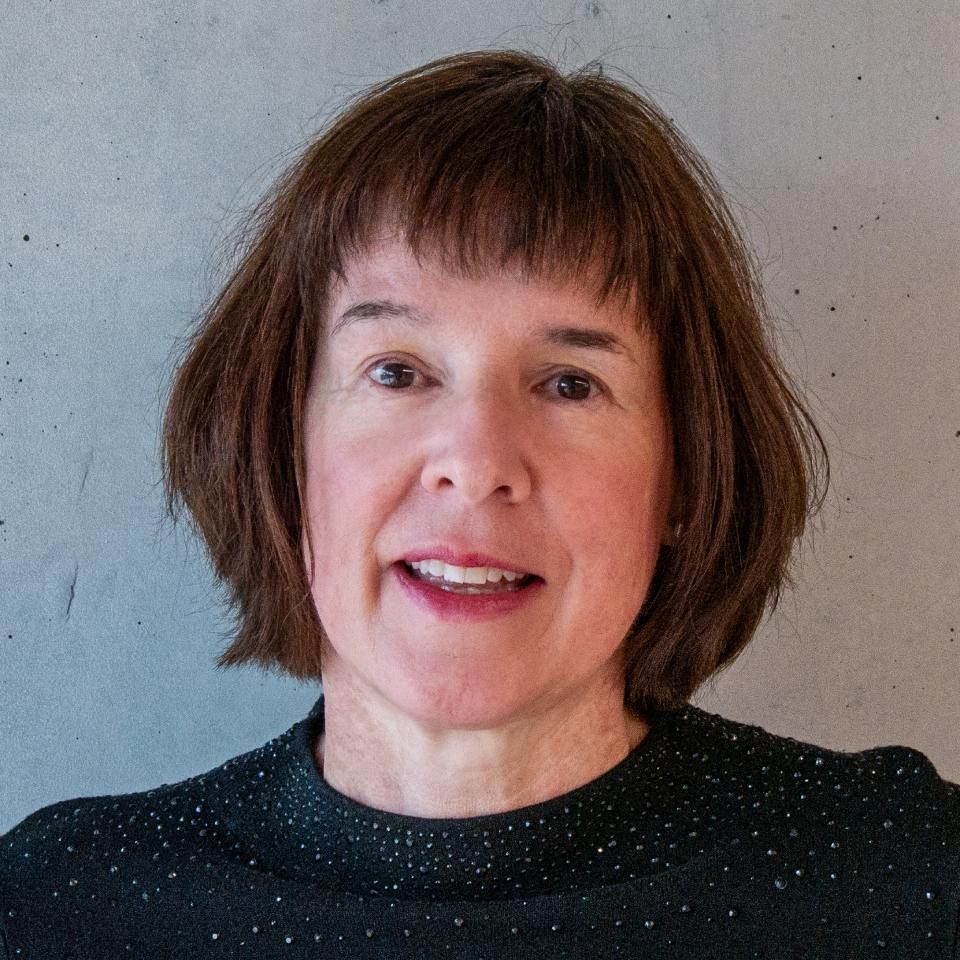 Porträt einer Person mit kurzen braunen Haaren und einem dunkel gesprenkelten Oberteil vor grauem Hintergrund (Kopf und Schultern).