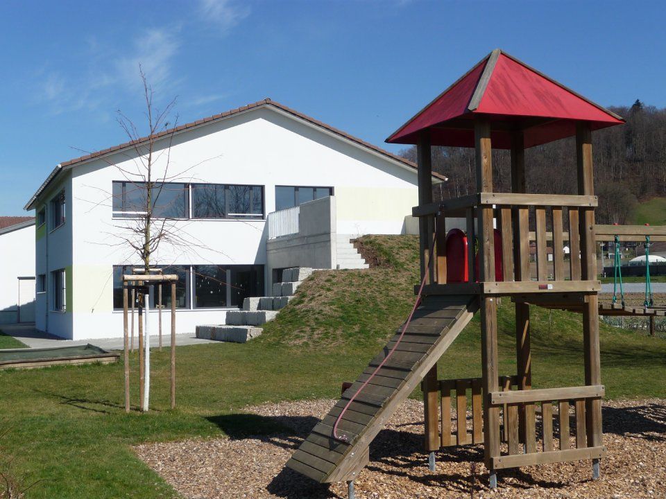 Spielplatzturm mit rotem Dach auf einer Rasenfläche neben einem weissen Gebäude unter klarem, blauem Himmel
