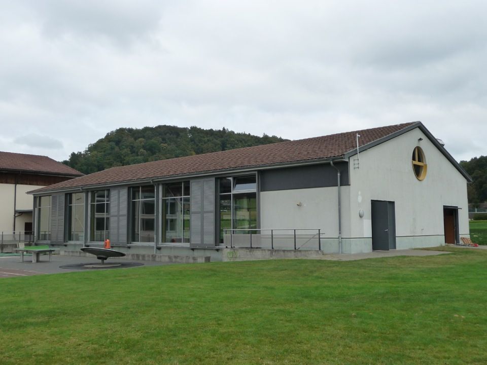 Eingeschossiges weisses Gebäude mit rotem Dach und grossen Fenstern neben einer grasbewachsenen Wiese unter bewölktem Himmel.