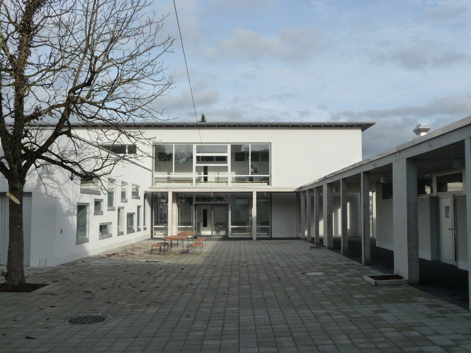 Innenhof eines weissen Gebäudes mit gepflasterter Terrasse, Fenstern und einem Baum im Vordergrund