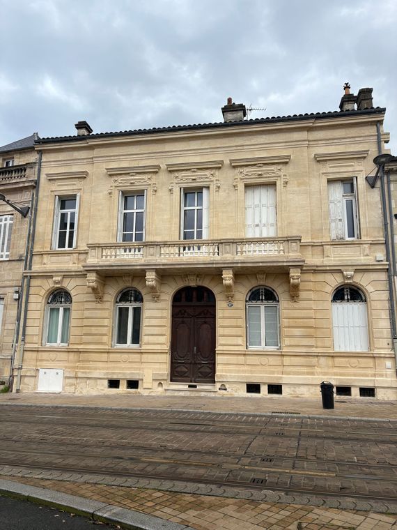 Bâtiment beige avec fenêtres et porte blanches, cyprès et marches en pierre menant à l'entrée.