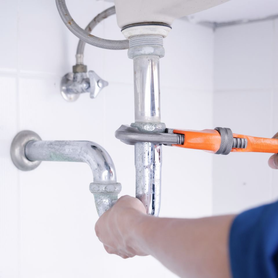 Un plombier utilise une clé pour réparer un tuyau argenté sous un évier blanc.