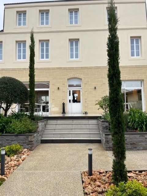 Bâtiment beige avec fenêtres et porte blanches, cyprès et marches en pierre menant à l'entrée.