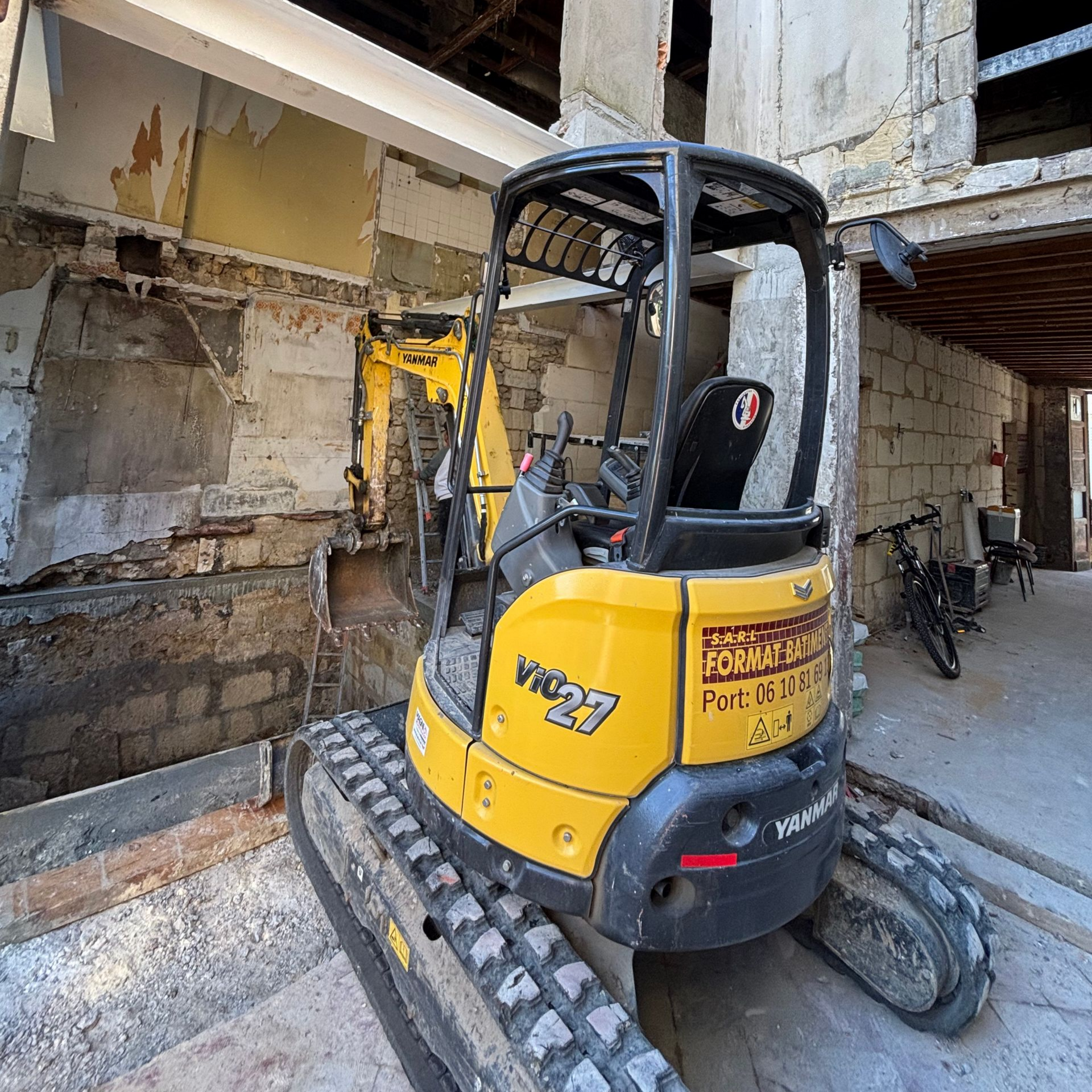 Une mini-pelle  jaune est garée à l'intérieur d'un bâtiment en cours de démolition, près d'un mur partiellement exposé.