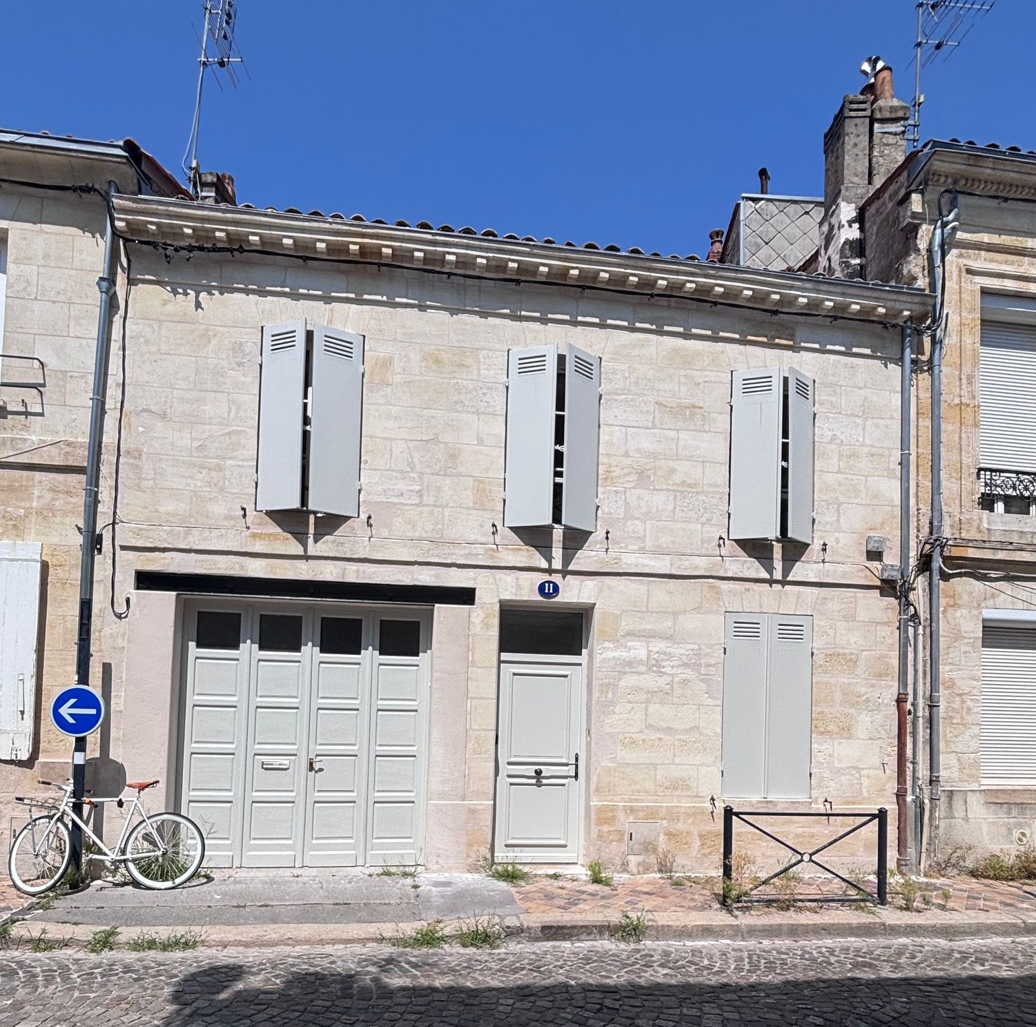 Extérieur d'une maison aux murs gris clair, au toit brun et aux volets blancs fermés.