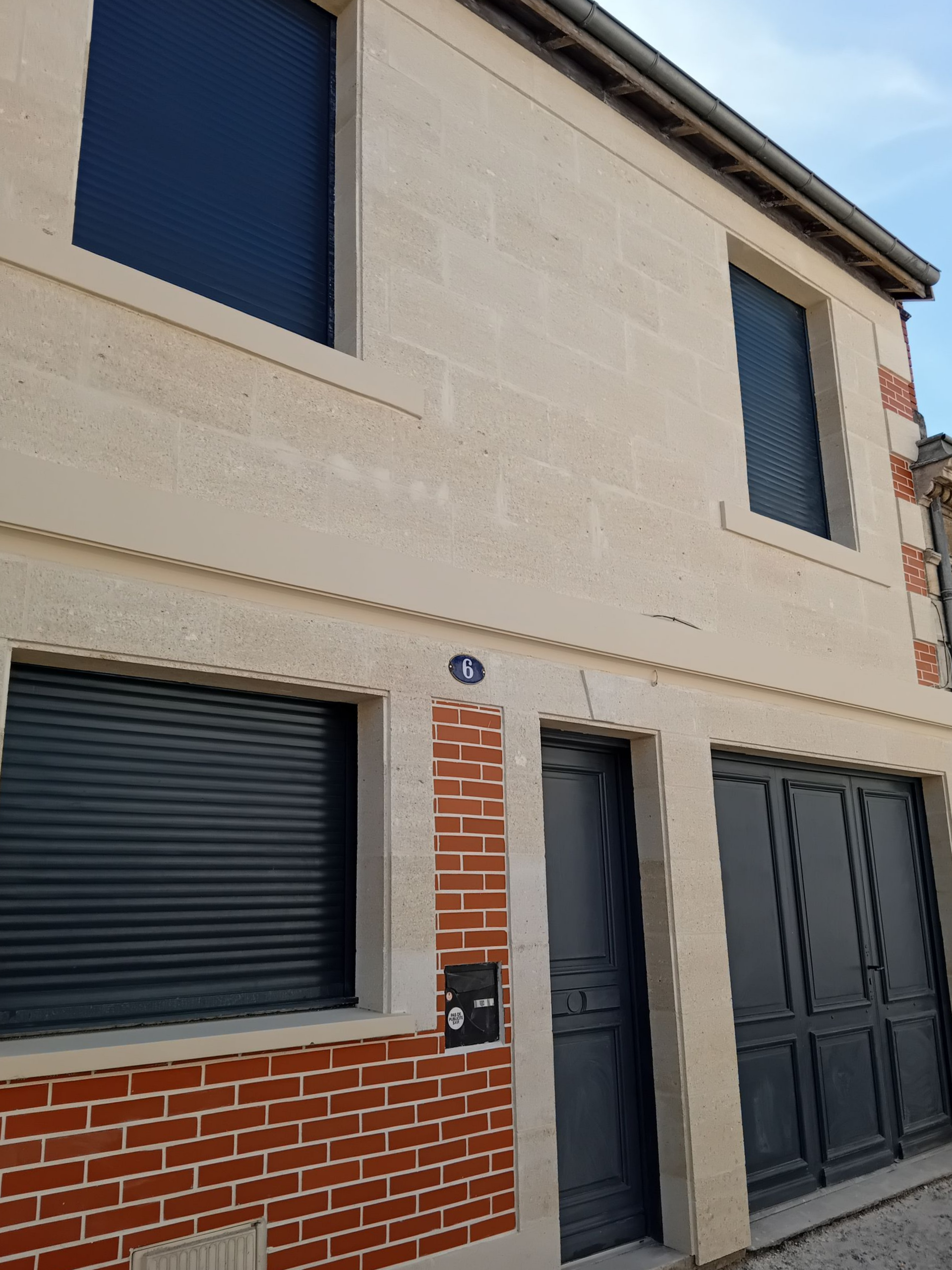 Bâtiment de deux étages à la façade beige et couverte en partie de briques ; stores bleu foncé. Porte et portail noirs.