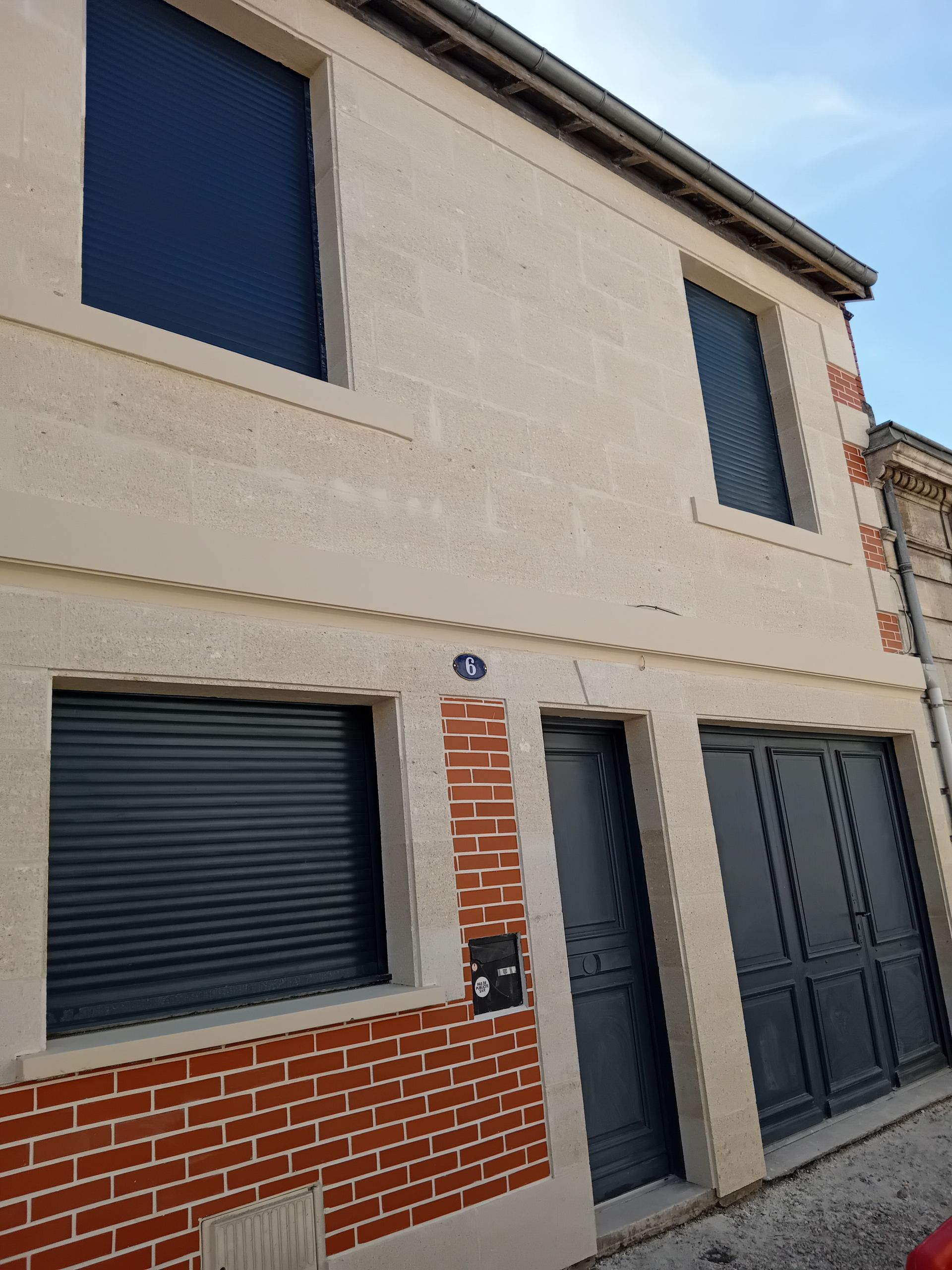 Un bâtiment de deux étages avec une façade en stuc clair, des accents de briques rouges et des volets et portes gris foncé.