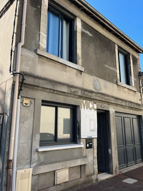 Un bâtiment de deux étages en stuc gris avec des encadrements de fenêtres gris foncé, une petite pancarte près de l'entrée et une porte de garage fermée.