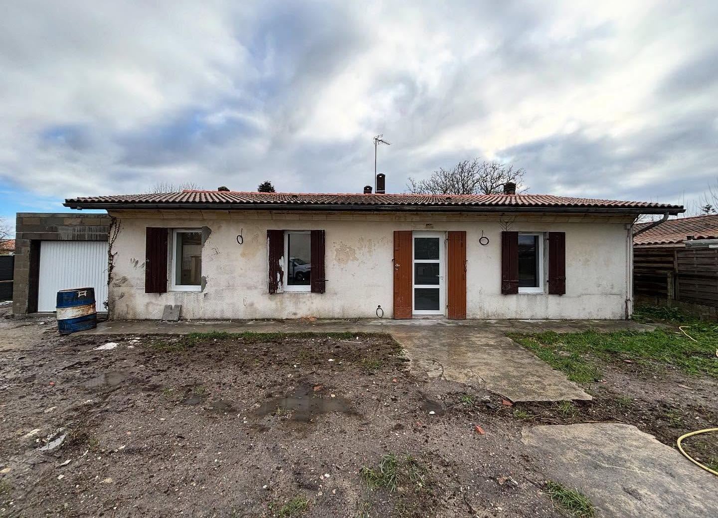 Maison blanche de plain-pied avec volets et porte marron. Allée en béton menant à l'entrée.
