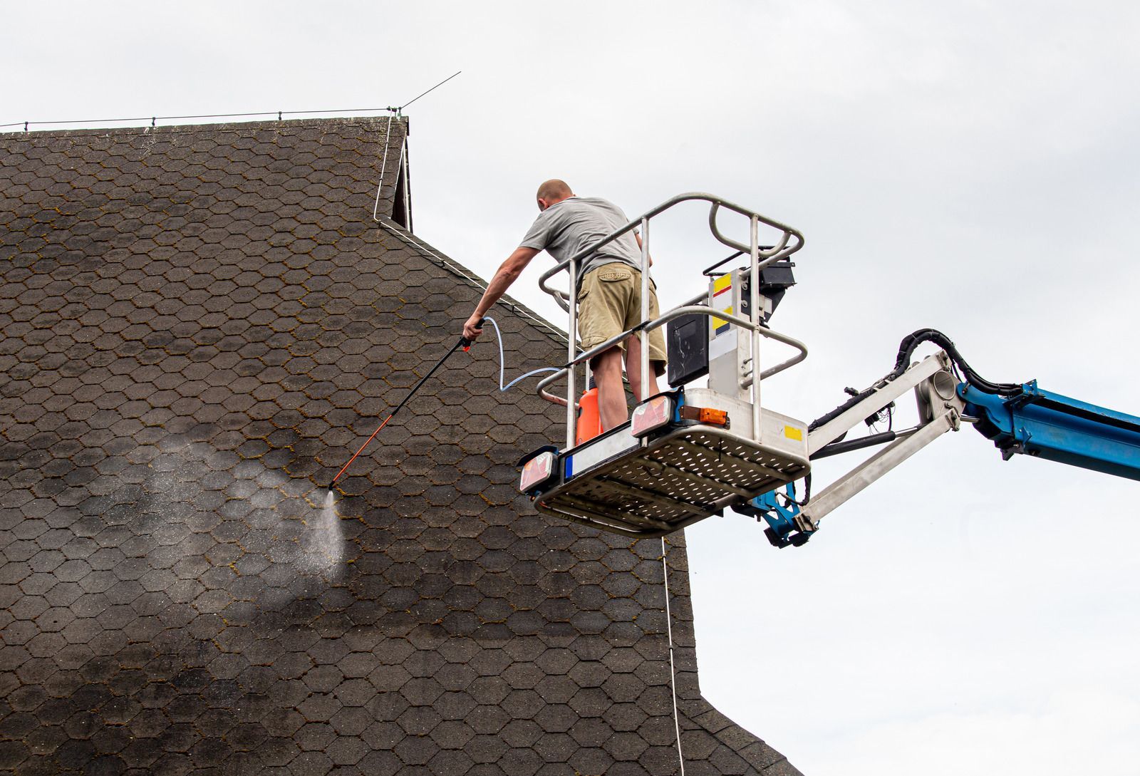 Worker on a blue boom lift repairing a shingled roof edge.